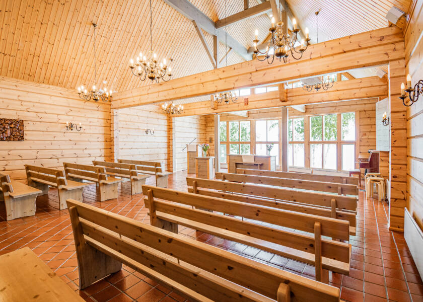Valoisa hirsinen sisätila, jossa kirkonpenkkejä, korkea katto ja kattokruunuja. Takaseinällä on ikkunoita. Lattia on oranssinpunaista laattaa.