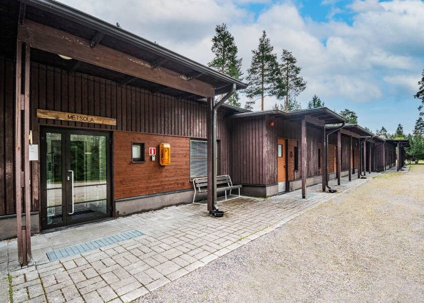 Ruskea pitkä matala rakennus, jossa monta etuovea. Isoimman oven päällä teksti: METSOLA