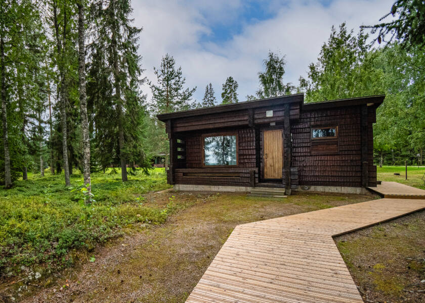 Tummanruskea pieni hirsirakennus metsäisessä ympäristössä.
