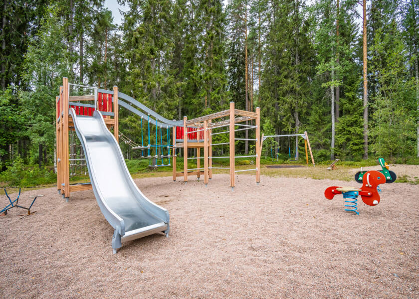Kiipeilyteline, liukumäki ja keinuja hiekkakentällä. Taustalla metsää.