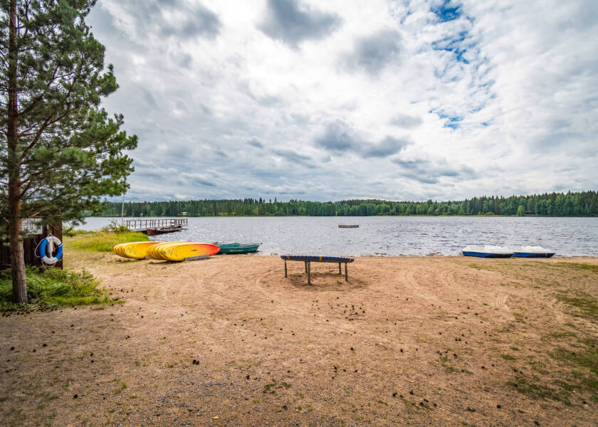 Kesäinen hiekkaranta ja järveä, rannalla suppilaitoja ja pari venettä.