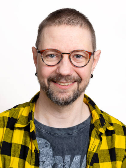 Henkilö. jolla lyhyet, tummat hiukset, parta, silmälasit ja kelta-mustaruudullinen paita.