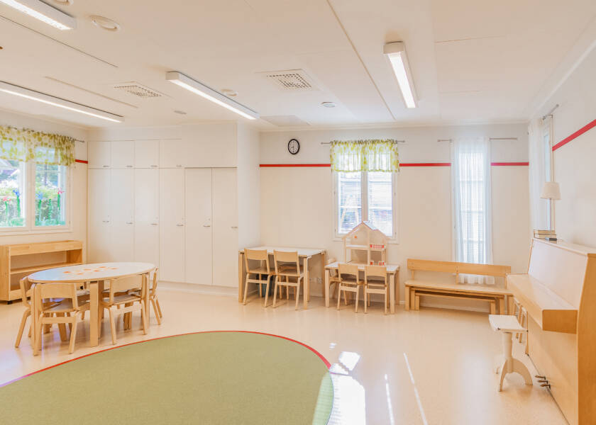 Valoisa tila jossa kiinteitöä valkoisia kaappeja, pari pöytäryhmää, piano ja penkki. Lattiatilaa jossa pyöreä vihreä matto.