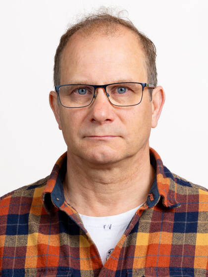 Henkilö jolla on lyhyet tummat hiukset, silmälasit ja värikäs, ruudullinen paita.