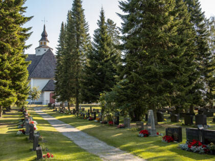 Hautausmaalla polku jonka molemmin puolin hautakiviä ja kukkia. Polun päässä kirkko. Isoja kuusia.