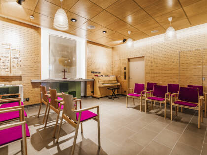 A bright room with birch-coloured ceiling and walls. On the back wall is an altar with a cross. On either side of the room are fuchsia-coloured pews.