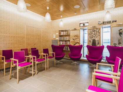 A bright room with birch-colored walls and ceiling, and a floor of brownish-gray tiles. Along the walls are chairs in shades of fuchsia.