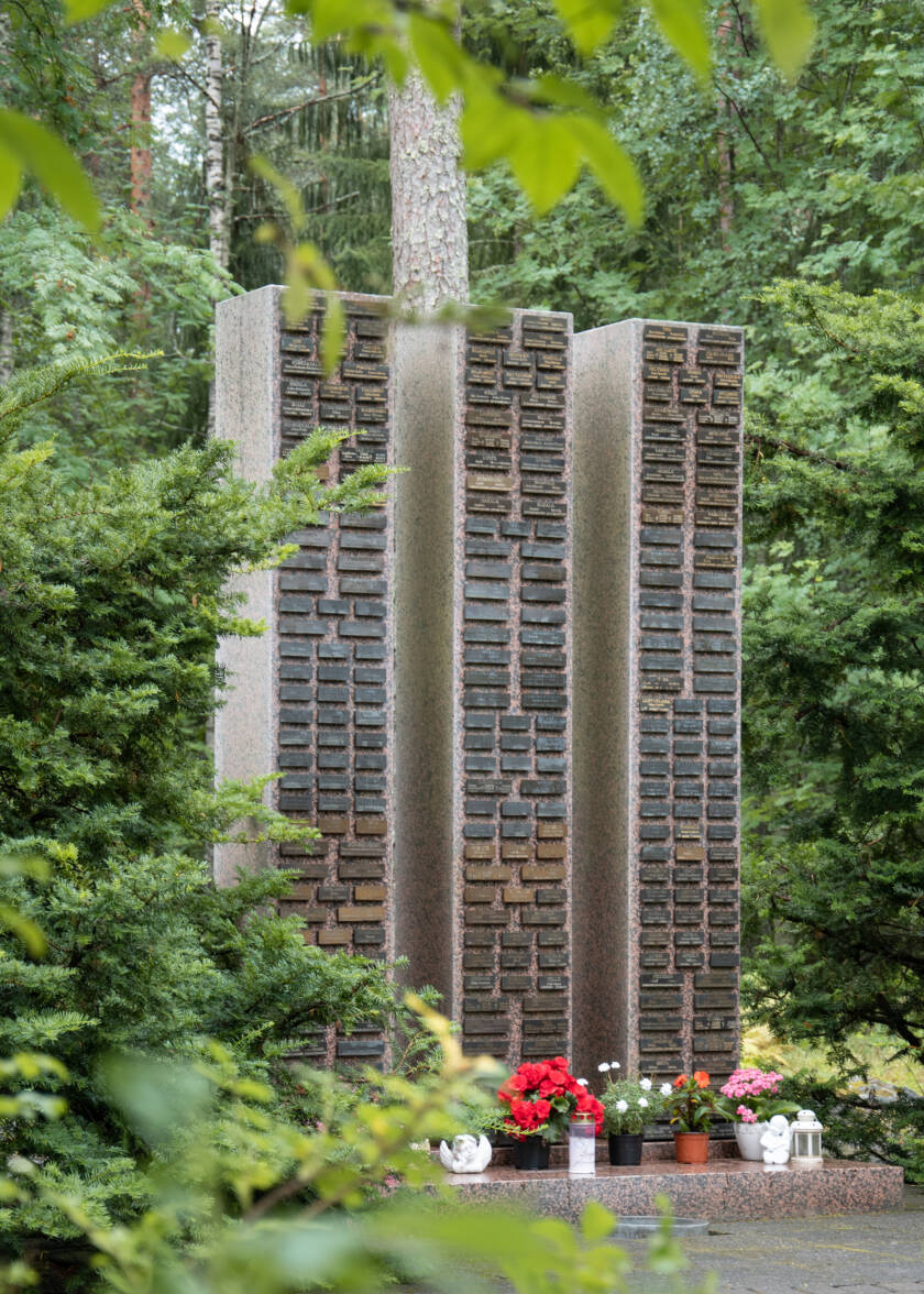 Kolme muistopylvästä hautausmaalla, edessä kukkia ruukuissa.