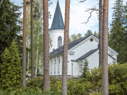 Puun runkoja joiden takaa näkyy valkoinen harmaakattoinen kirkko.