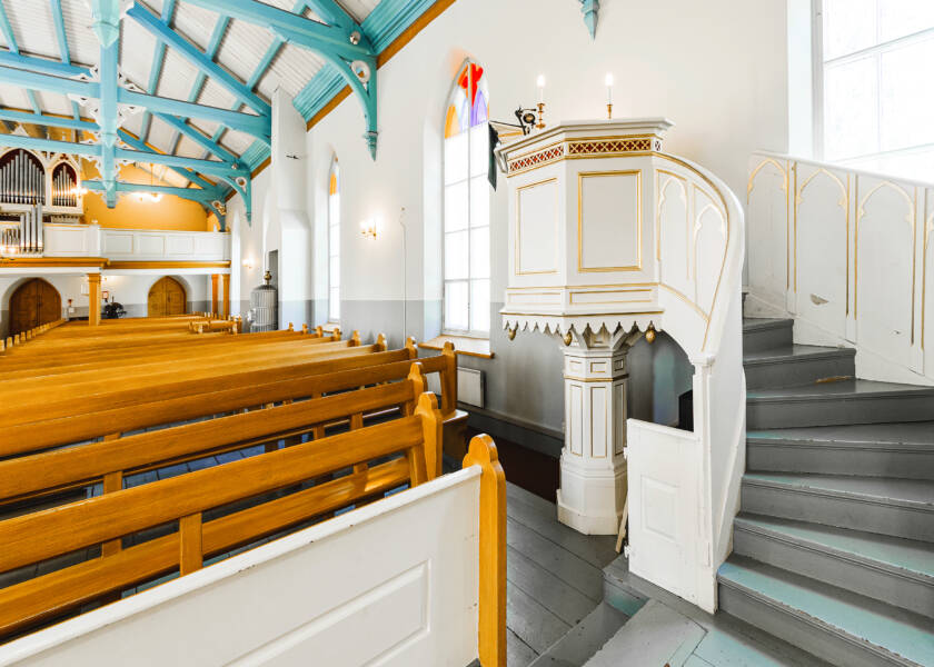 Oikealla valkoinen saarnastuoli kultaisilla ja punaisilla yksityiskohdilla ja vasemmalla kirkon puunväriset penkkirivit.