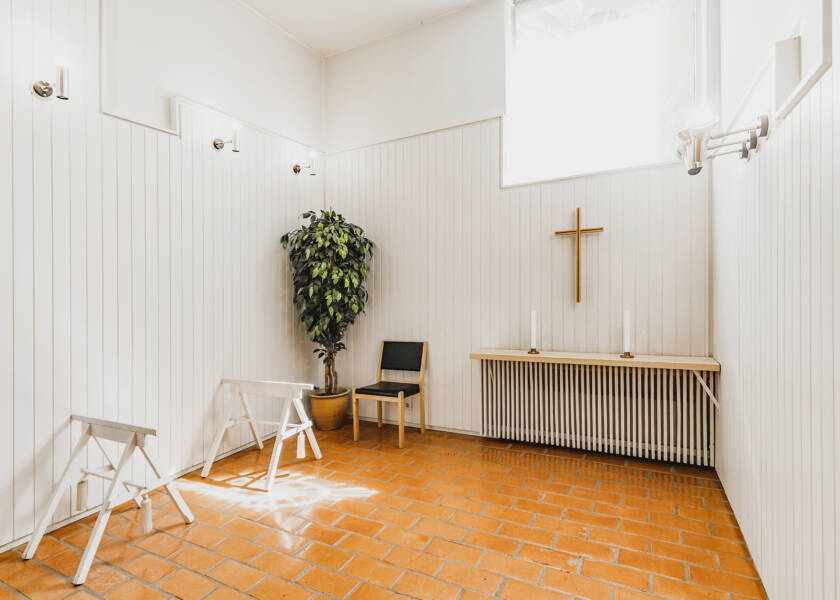 Valkoinen pieni tila, jossa pieni alttari kahdella kynttilällä. Seinällä risti. Nurkassa huonekasvi. Lattia punaista tiiltä. Seinän vieressä tukit hauta-arkkua varten.