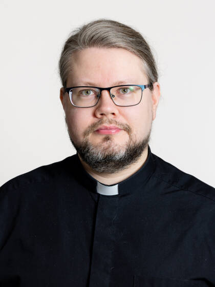 Kasvokuva henkilöstä jolla on hiukset takana kiinni, silmälasit, viikset ja partaa sekä papinkaulus.