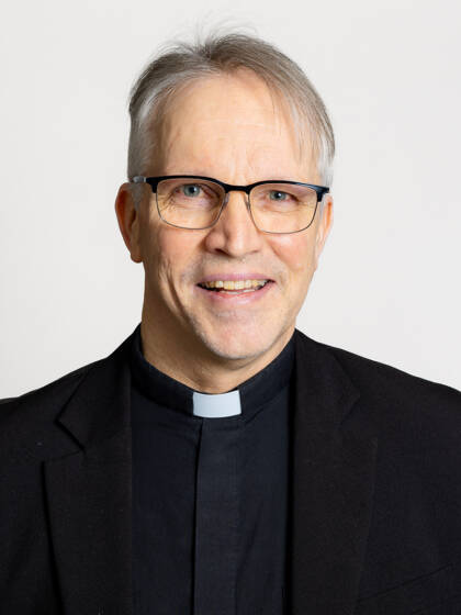 Close-up portrait of a person with short hair, glasses, a clerical collar, and a suit jacket.
