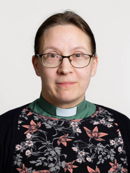 Kasvokuva henkilöstä jolla on hiukset kiinni takana, silmälasit ja papinkaulus vihreällä paidalla.