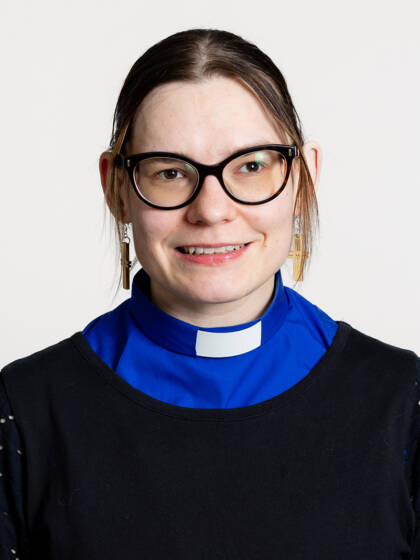 Kasvokuva henkilöstä jonka hiukset ovat kiinni takana, paksusankaiset mustat silmälasit sekä papinkaulus sinisessä paidassa.