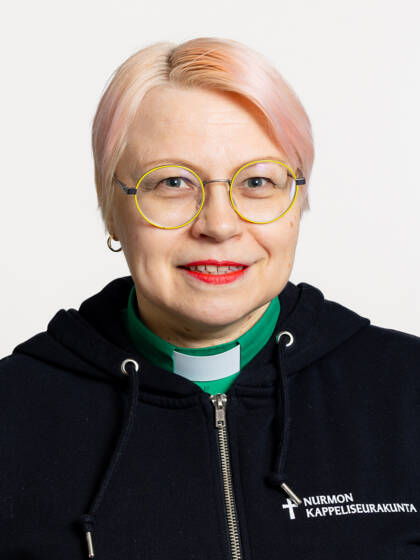 Henkilö jolla on vaaleanpunaiset lyhyet hiukset, keltasankaiset silmälasit, punaista huulipunaa sekä papinkaulus vihreässä paidassa.