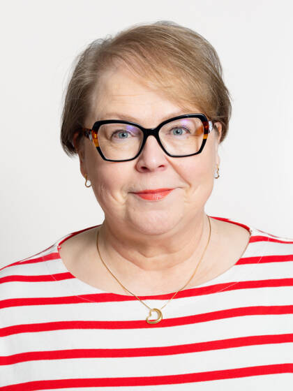 Kasvokuva henkilöstä jolla on lyhyet vaaleat hiukset sekö paksusankaiset silmälasit, punaista huulipunaa sekä punavalkoraidallinen paita.