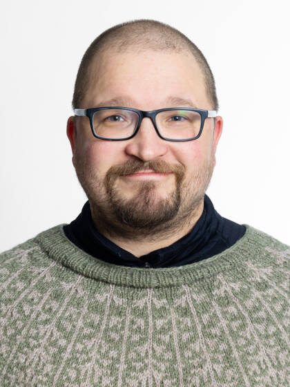 Kasvokuva henkilöstä jolla on siili tukka ja mustasankaiset silmälasit sekä lyhyttä partaa ja viikset.