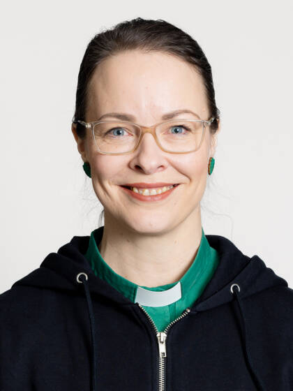 Kasvokuva henkilöstä jolla on tummat hiukset kiinni takana ja papinkaulus vihreässä paidassa sekä silmälasit.