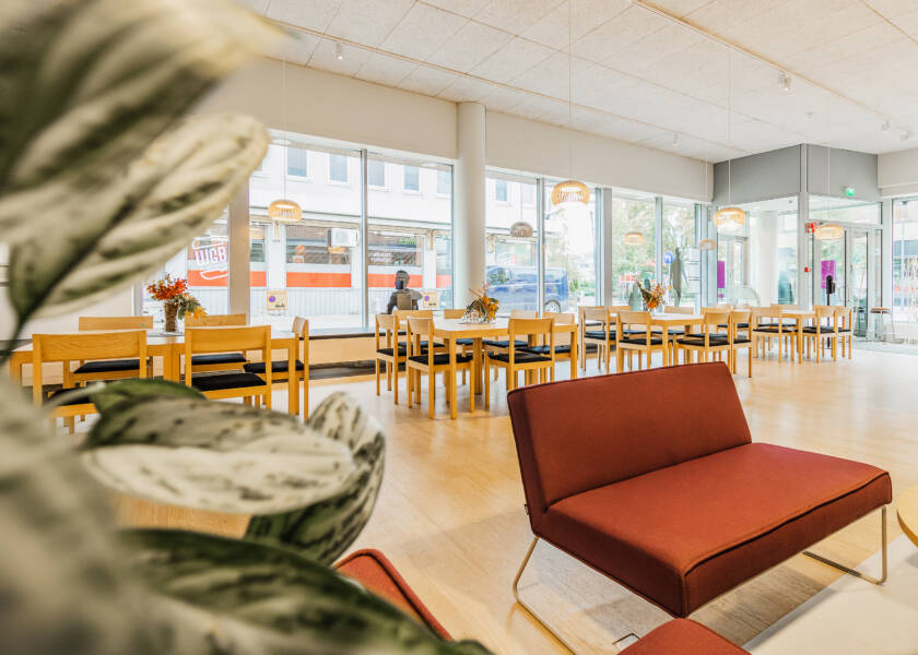 Valoisa tila jossa suuret ikkunat kadulle päin. Tilassa pöytäryhmiä. Etualalla punainen tuoli ja viherkasvi.
