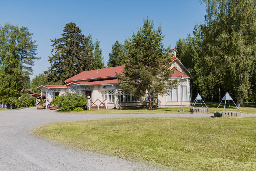 Pihatietä jossa punakattoinen rakennus, rakennuksen ympärillä puita ja pensaita.