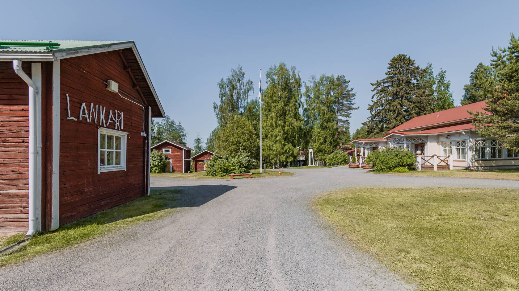 Pihatie jonka vasemmalla puolella punainen hirsirakennus jonka seinässä lukee LANKARI. Oikealla puolella punakattoinen talo, taustalla vihertäviä puita.
