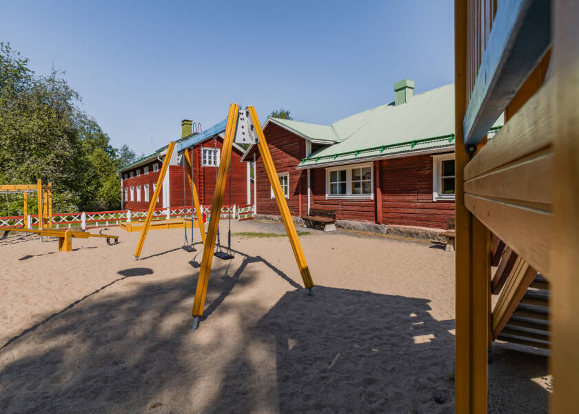 Hiekkapohjainen leikkikenttä jossa keinuja ja kiipeilyteline. Taustalla kaksi punaista hirsirakennusta.