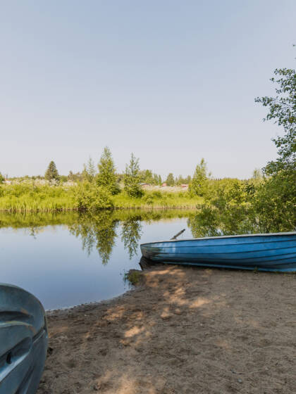 Metsäinen ranta ja järveä. Hiekalla sininen soutuvene.