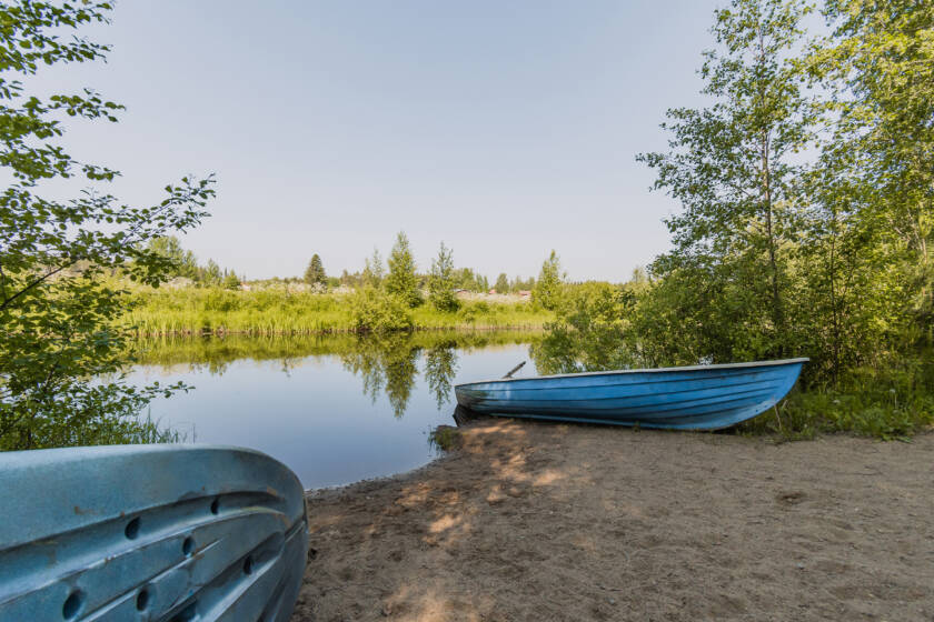 Metsäinen ranta ja järveä. Hiekalla sininen soutuvene.