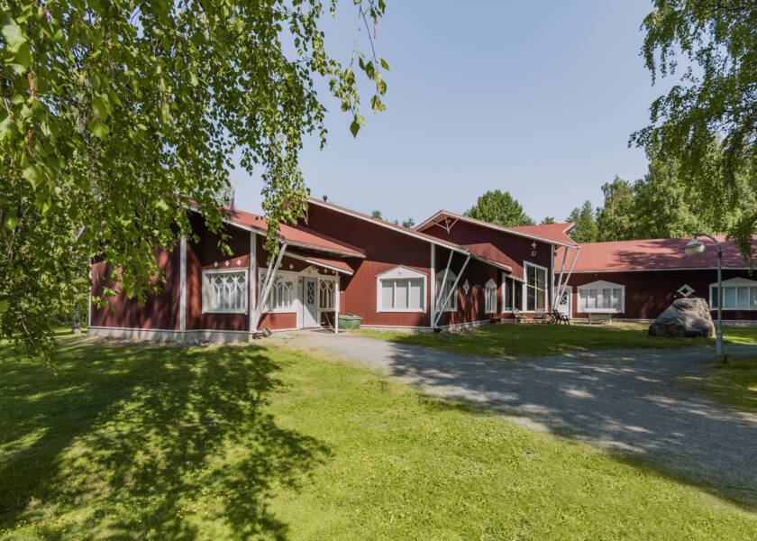 Punainen rakennus jossa kattoa neljällä eri tasolla. Ympärillä vihertäviä koivuja.