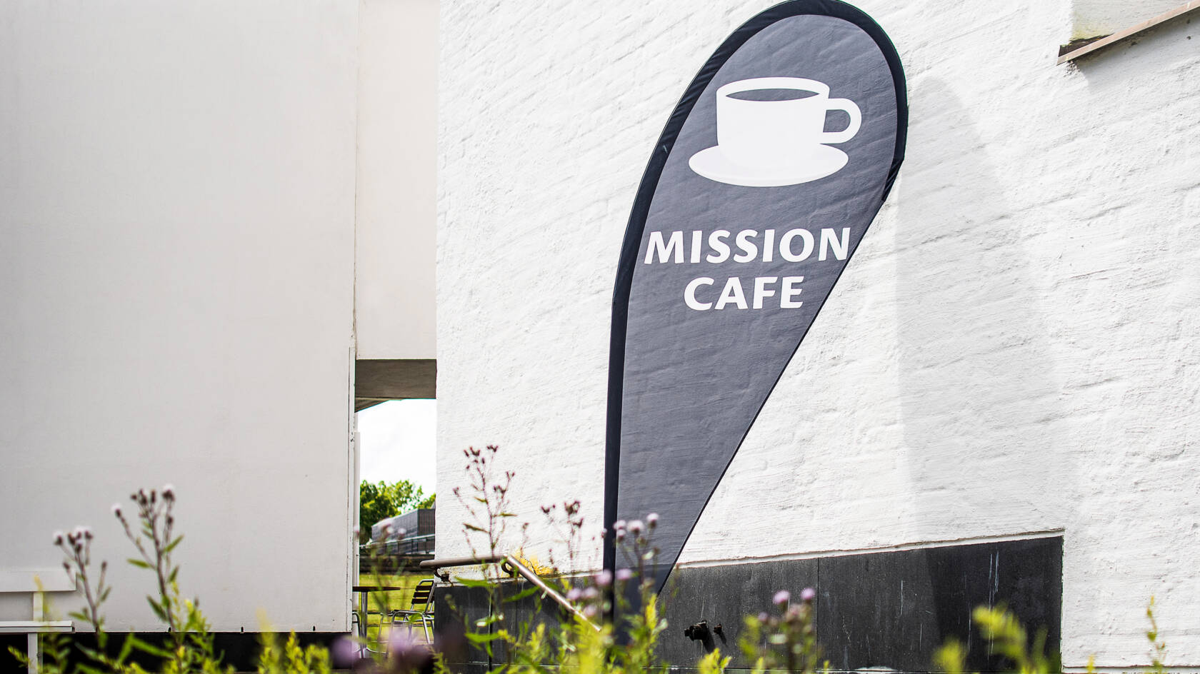Valkoista tiilirakennusta vasten musta pisaranmuotoinen lippu jossa valkoinen kahvikuppi ja teksti MISSION CAFE.