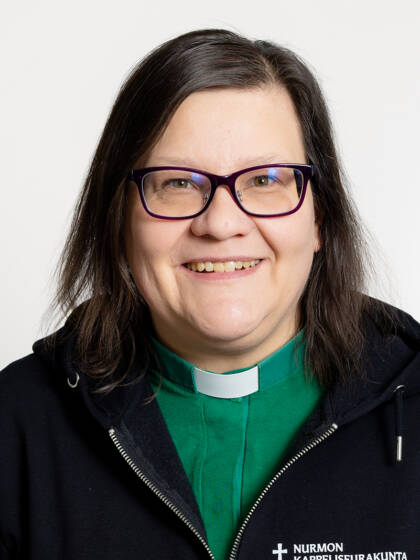 Henkilö jolla on on olkapäille ulottuvat tummat hiukset, silmälasit sekä papinkaulus vihreällä paidalla.