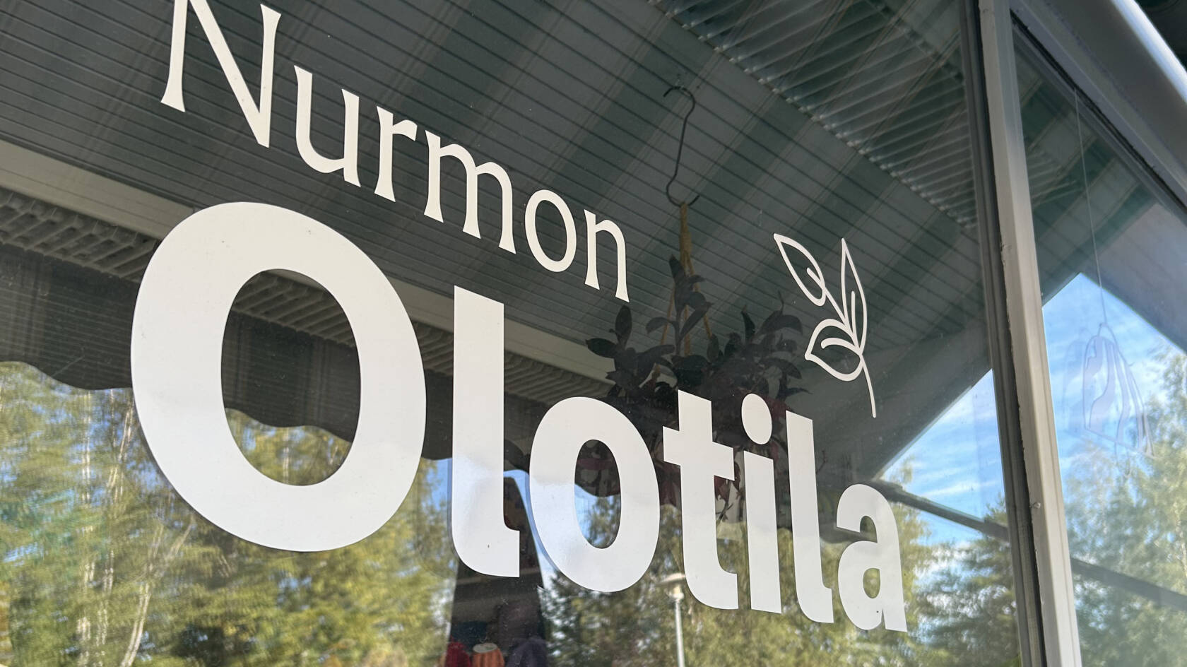 Ikkuna johon teipattu teksti: Nurmon Olotila. Ikkunasta heijastuu puita ja rakennuksia.