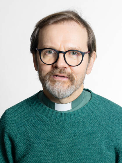 Kasvokuva henkilöstä jolla on mustasankaiset silmälasit, lyhyet hiukset sekä papinkaulus vihreässä paidassa.