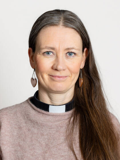 Henkilö jolla on pitkät ruskeat hiukset, sydänkorvakorut ja papinkaulas sekä vaalea neule.