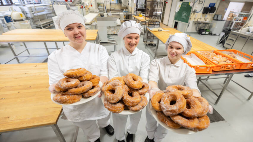 Kolme henkilöä valkoisissa kokin asuissa ja kokin hatussa pitävät munkkilautasia käsissään. Taustalla suurtalouskeittiö.