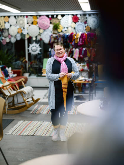 A person in Nurmo Living Room holding a knitting project, with colourful handicrafts in the background.