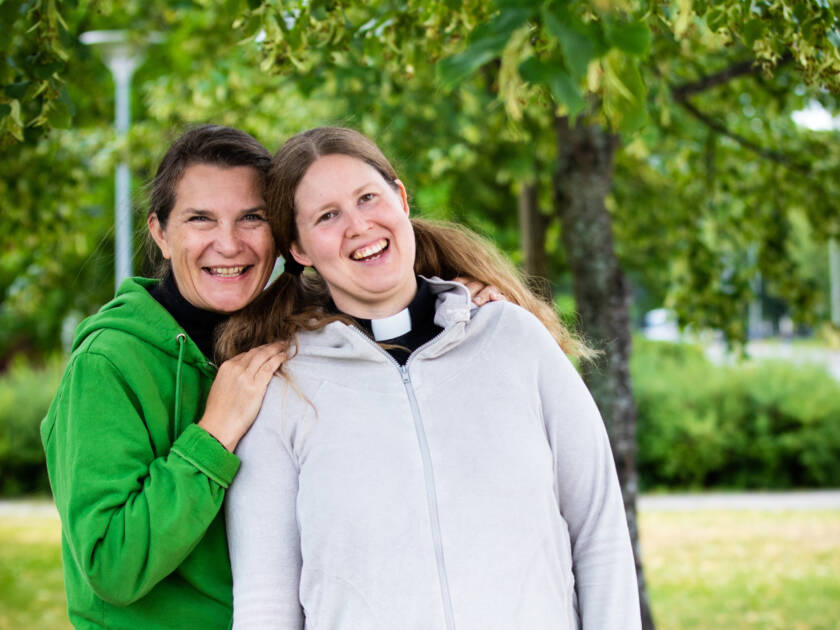 Kaksi ihmistä vihreiden puiden keskellä. Toinen pitää käsiään toisen harteilla.
