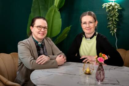 Diakoniatyöntekijä ja pappi istuvat pyöreän pöydän ääressä. Pöydällä pinkkejä kukkia ja kynttilä. Taustalla vihreä seinä ja viherkasvi.