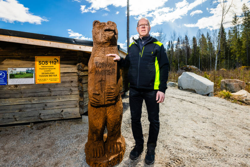 Puusta veistetty karhupatsas ja henkilö vierekkäin. Taustalla laavu sekä metsäistä maisemaa.