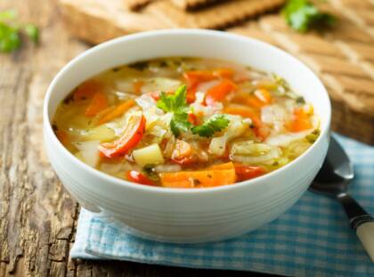 A bowl of clear vegetable soup on a wooden table, with a spoon beside the bowl.