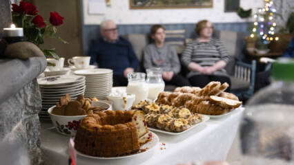 Kuvan etualalla pöytä, jolle katettu kahvia ja leivonnaisia, taustalla istuu ihmisiä.