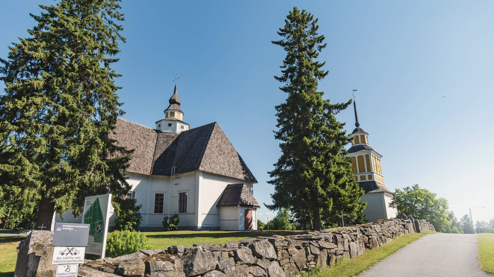 Vaalea ruskeakattoinen kirkkorakennus, etualalla kiviaitaa.