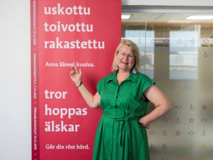 Ihminen seisoo punaisen värisen seurakuntavaalimainoksen vieressä vihreässä mekossa.