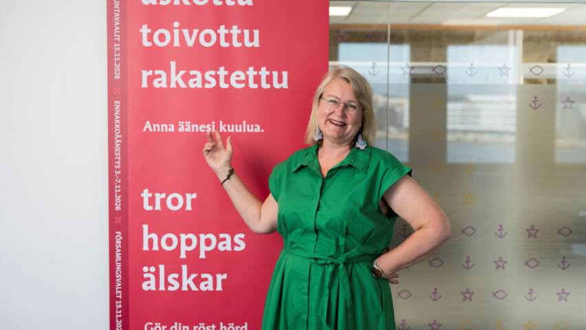 Ihminen seisoo punaisen värisen seurakuntavaalimainoksen vieressä vihreässä mekossa.