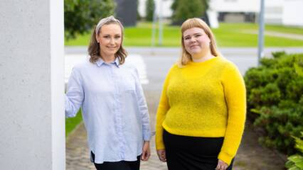Kaksi ihmistä seisovat vierekkäin pylvään vieressä. Toisella henkilöllä on sinertävä paita, sekä musta alaosa ja hän nojaa pylvääseen. Toisella henkilöllä on keltainen paita ja musta alaosa. Taustalla pensaita ja puita.