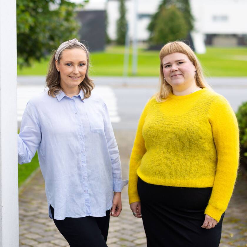 Kaksi ihmistä seisovat vierekkäin pylvään vieressä. Toisella henkilöllä on sinertävä paita, sekä musta alaosa ja hän nojaa pylvääseen. Toisella henkilöllä on keltainen paita ja musta alaosa. Taustalla pensaita ja puita.