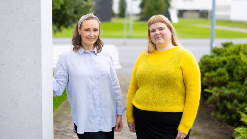 Kaksi ihmistä seisovat vierekkäin pylvään vieressä. Toisella henkilöllä on sinertävä paita, sekä musta alaosa ja hän nojaa pylvääseen. Toisella henkilöllä on keltainen paita ja musta alaosa. Taustalla pensaita ja puita.
